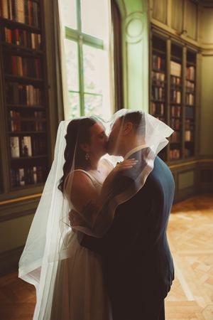 Bride and groom kissing under a flowing veil in an elegant library in the Meridian House DC with bookshelves in the background.