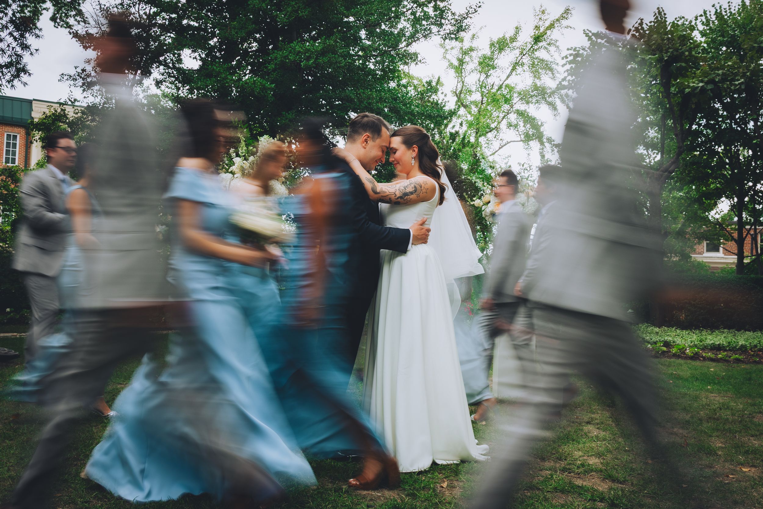 Bride and groom share a romantic moment outdoors while wedding party members blur past them in motion.