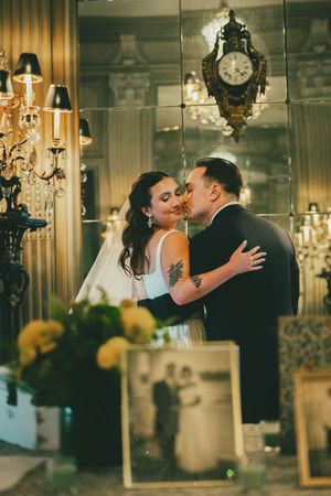 Bride and groom share a romantic kiss in an elegant mirrored venue with chandeliers and vintage clock decor.