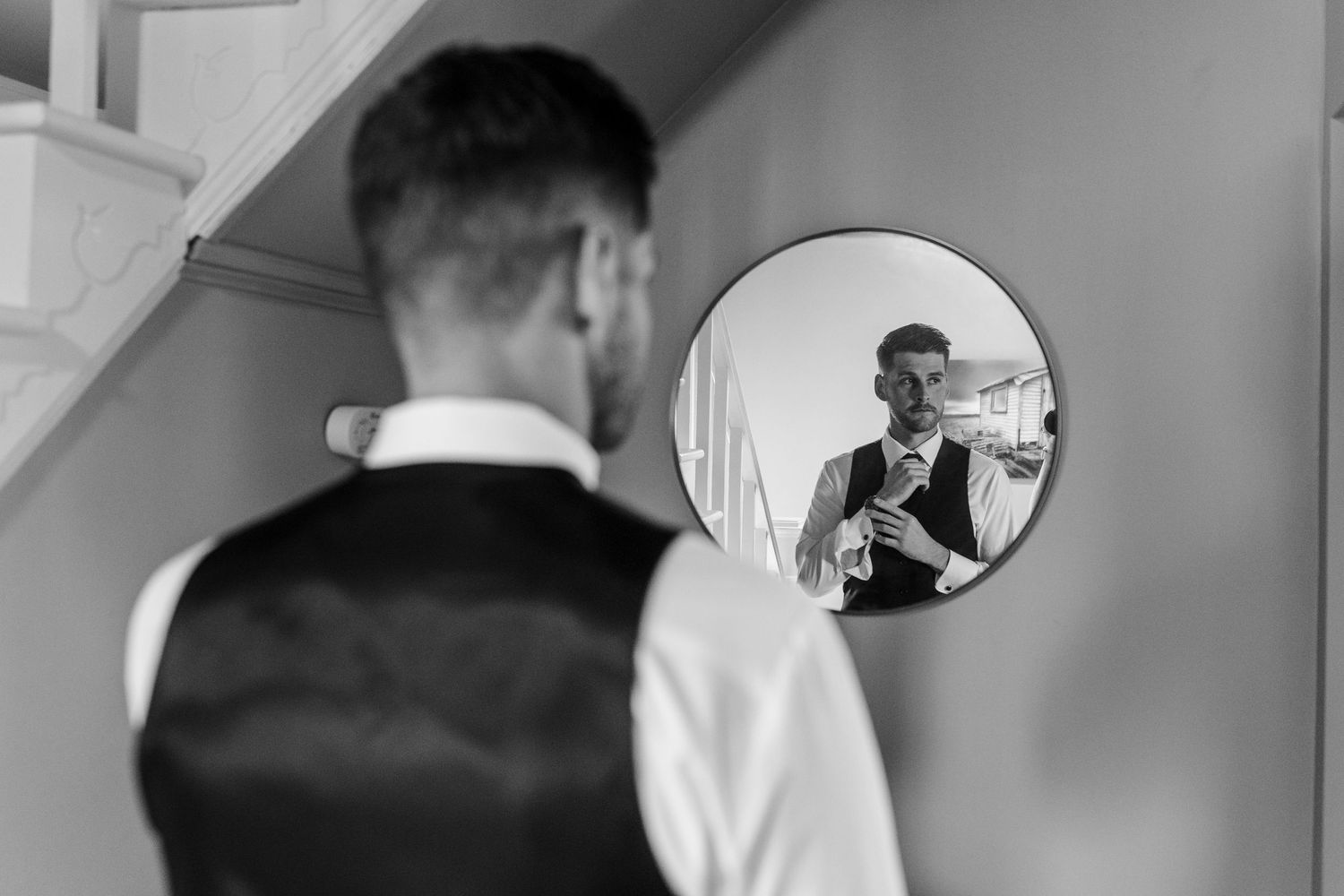 Black and white photo of a groom adjusting his tie in a round mirror while getting ready for his wedding day.