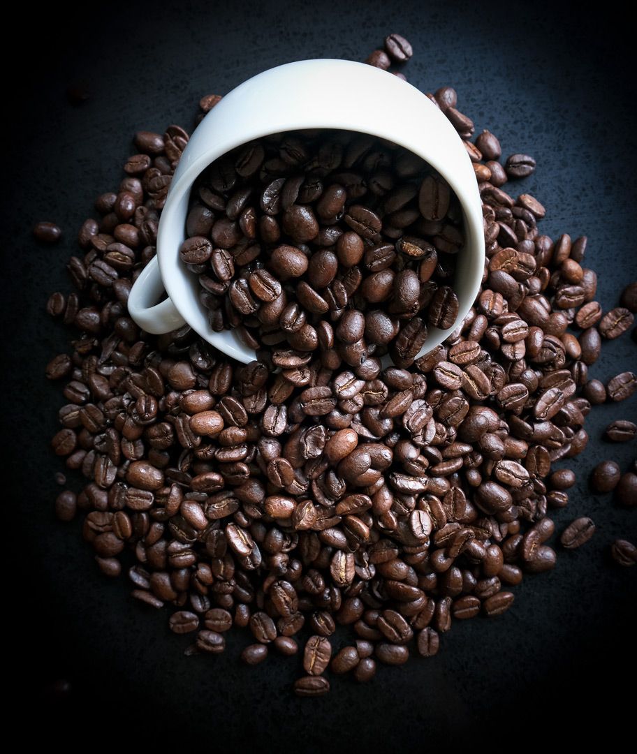 A white ceramic cup tipped over spilling roasted coffee beans onto a dark surface.