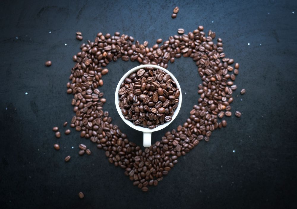 A white coffee cup filled with roasted coffee beans arranged in a heart shape on a dark slate background.