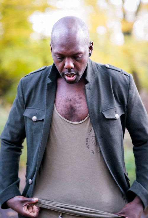 Bald Black man in olive jacket and tan tank top looking down outdoors with blurred green foliage background.
