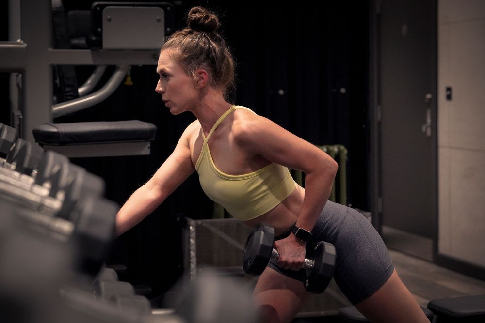 Woman performing dumbbell rows in a gym, wearing a yellow sports bra and gray shorts, showcasing strength training form.