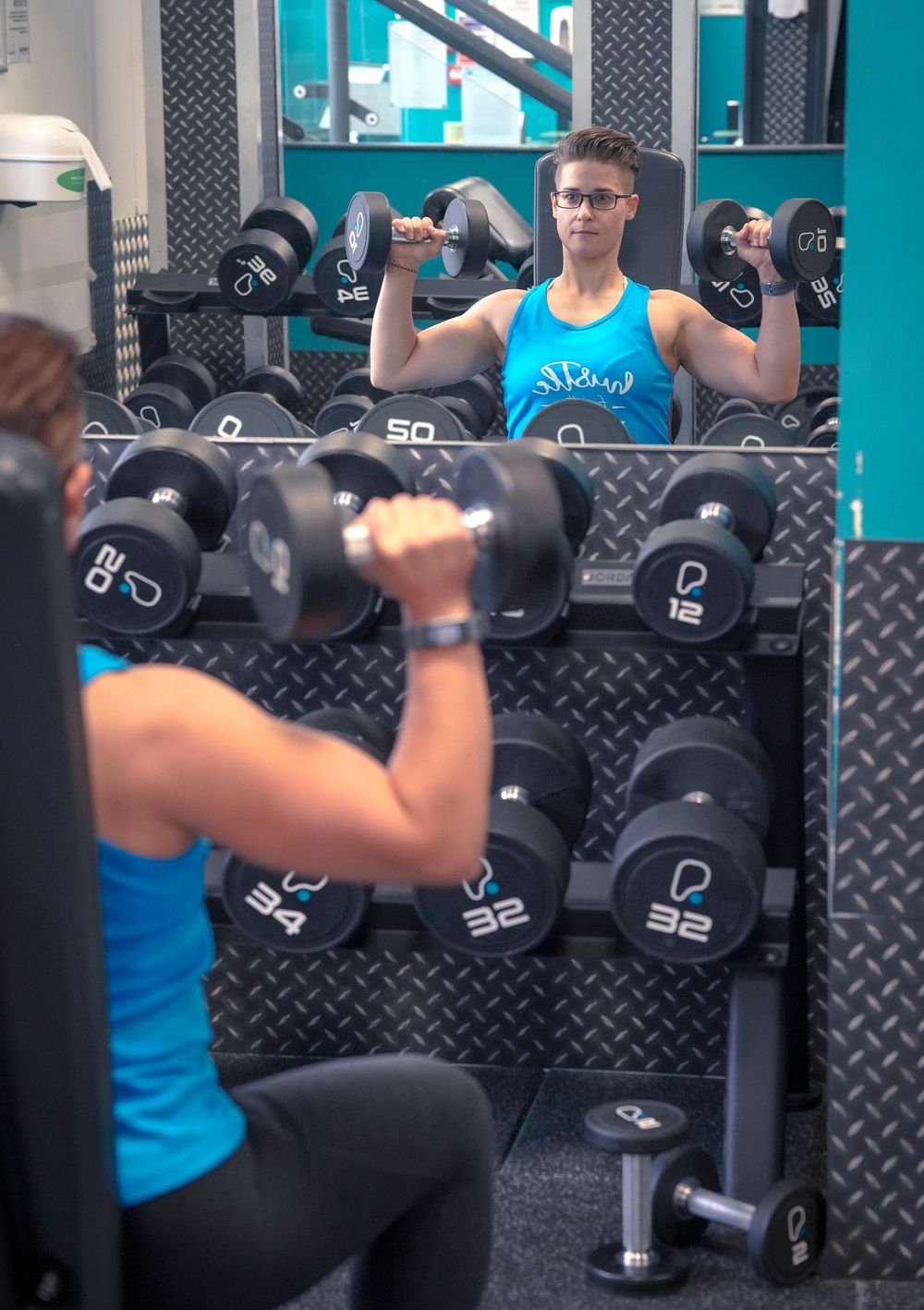 Two people lifting dumbbells in a gym, with a rack of weights visible in the background.