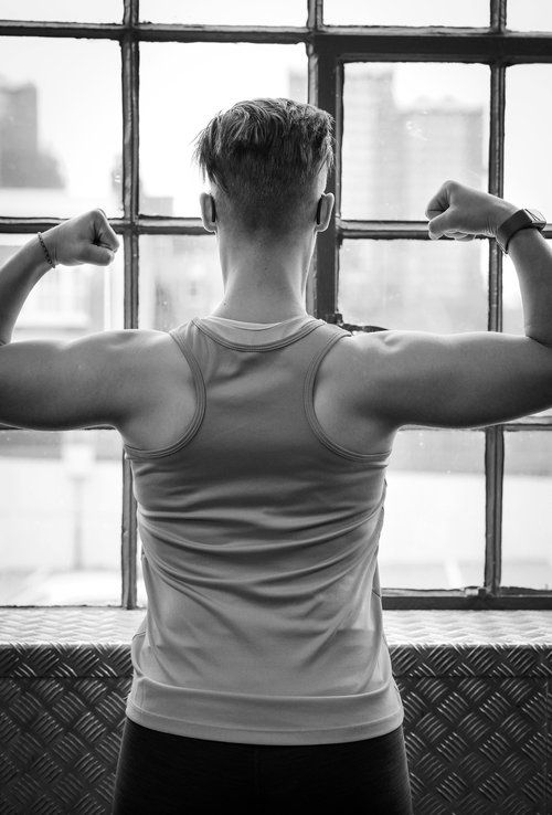 Muscular man in white tank top flexing biceps from behind near industrial window in black and white photo.
