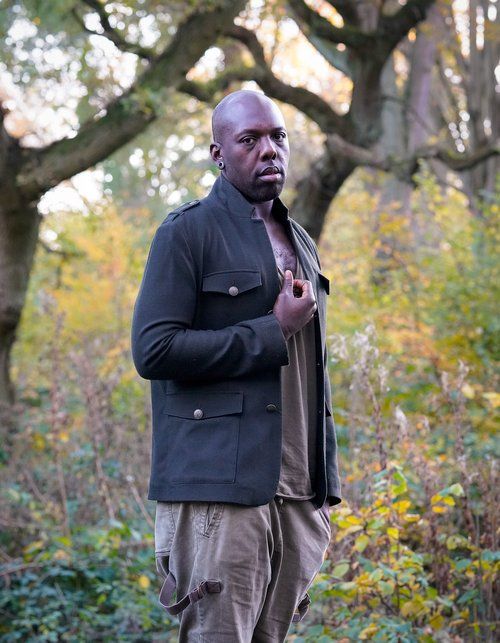 Bald man in dark jacket and cargo pants poses in autumn forest setting.