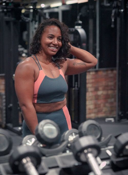 Smiling woman in gray and coral sports bra and leggings poses confidently in a gym near a rack of dumbbells.
