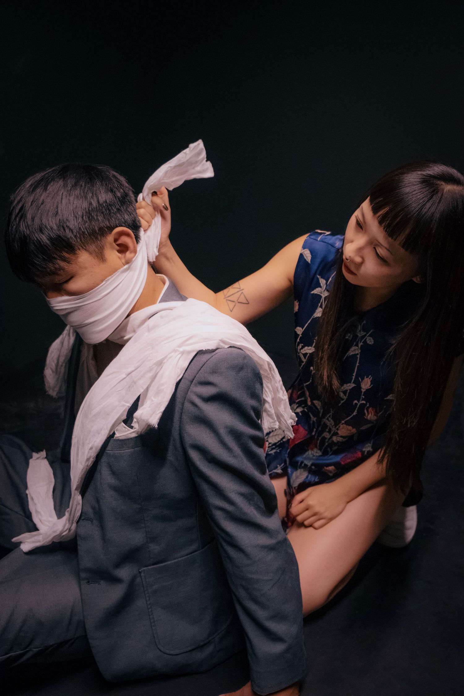 A woman in a blue floral dress wraps white fabric around a seated man in a gray suit against a dark background.
