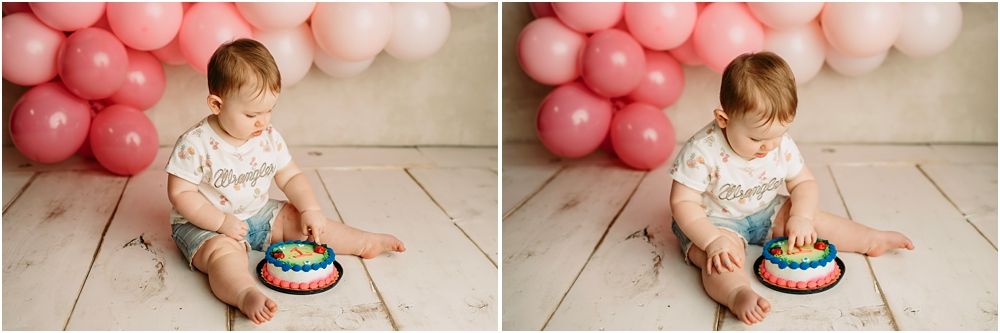 Baby girl in white outfit sits on floor with rainbow cake and pink balloons during first birthday cake smash photo session.