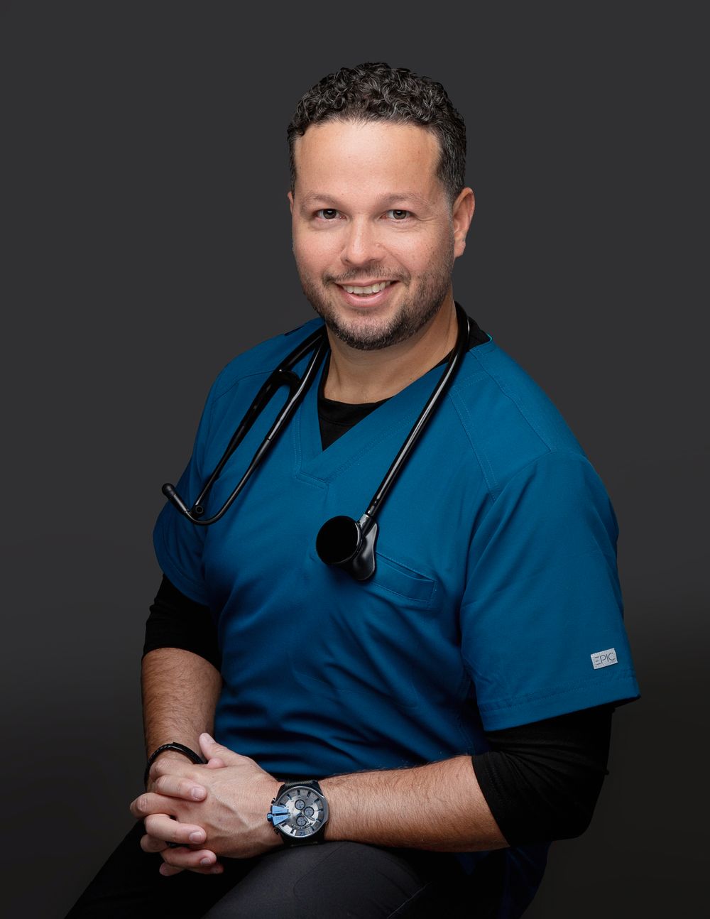 Doctor in navy scrubs with arms crossed on a dark backdrop, executive headshot