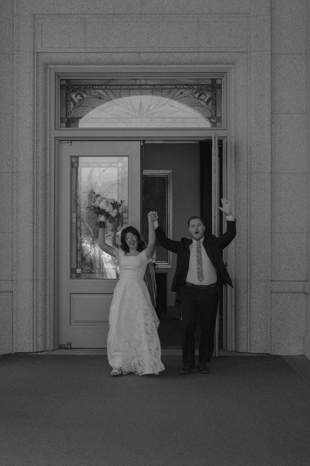 Joyful newlyweds raising hands in celebration as they exit through ornate church doors in black and white.