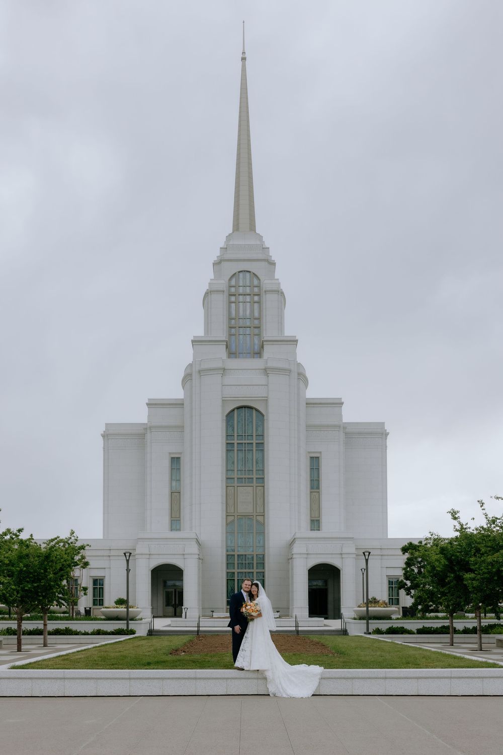 Bride and groom embrace in front of a white LDS temple with tall spire on a cloudy wedding day.
