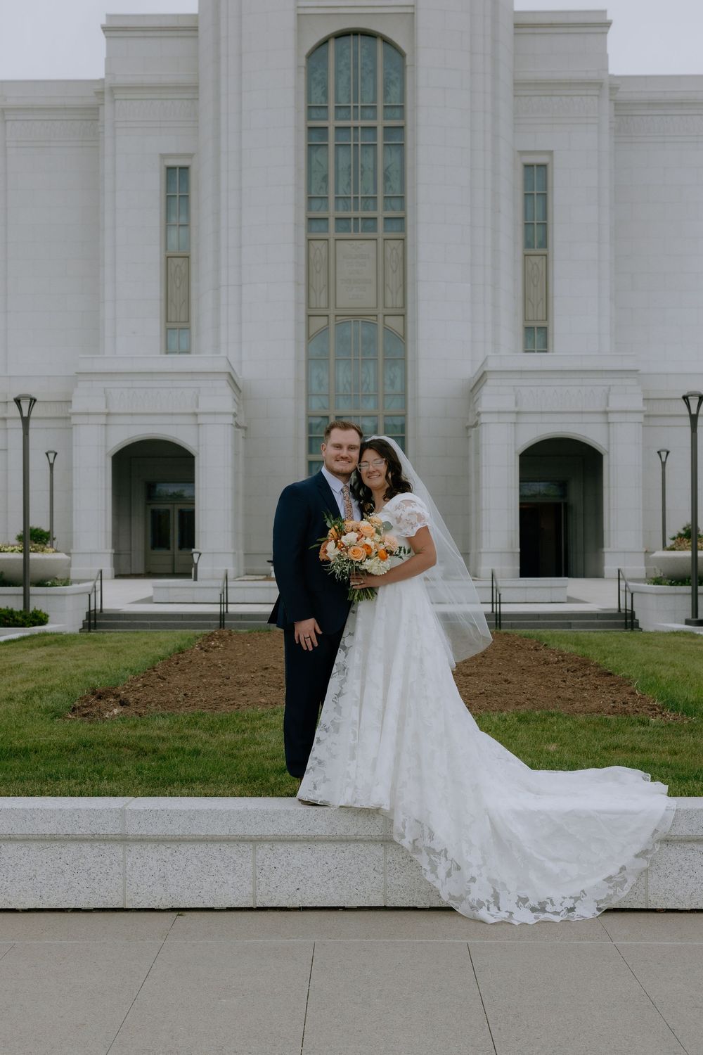 Bride and groom posing in front of a white LDS temple, bride holding orange bouquet in lace wedding gown.