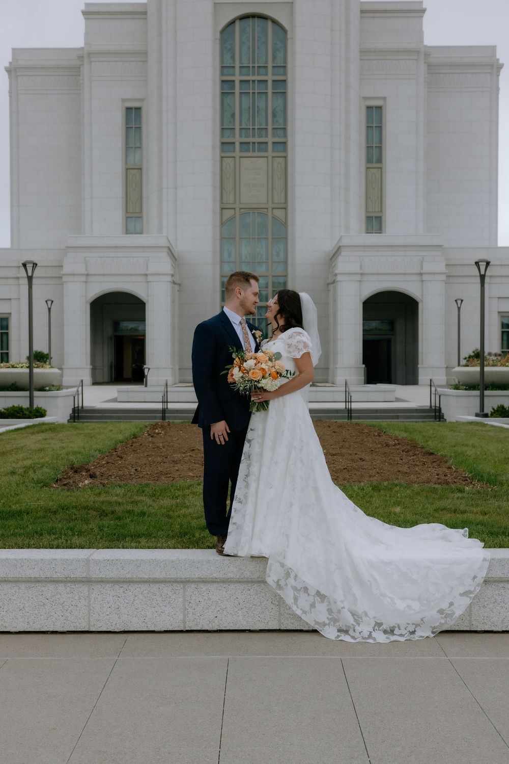 Bride and groom share tender moment before grand white LDS temple with lush green landscaping.
