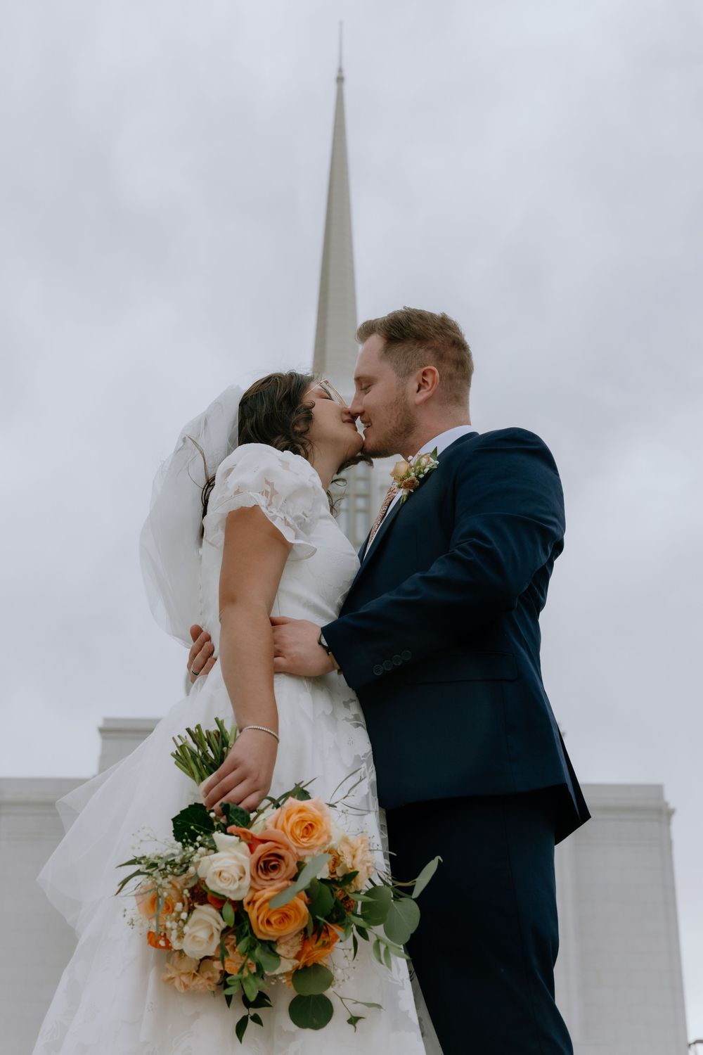 Bride and groom kissing in front of a church steeple, bride holding orange and white rose bouquet.