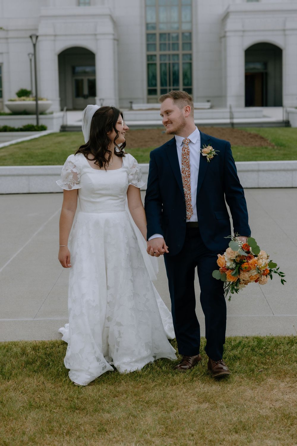 Bride and groom walk hand in hand smiling at each other outside a white wedding venue.