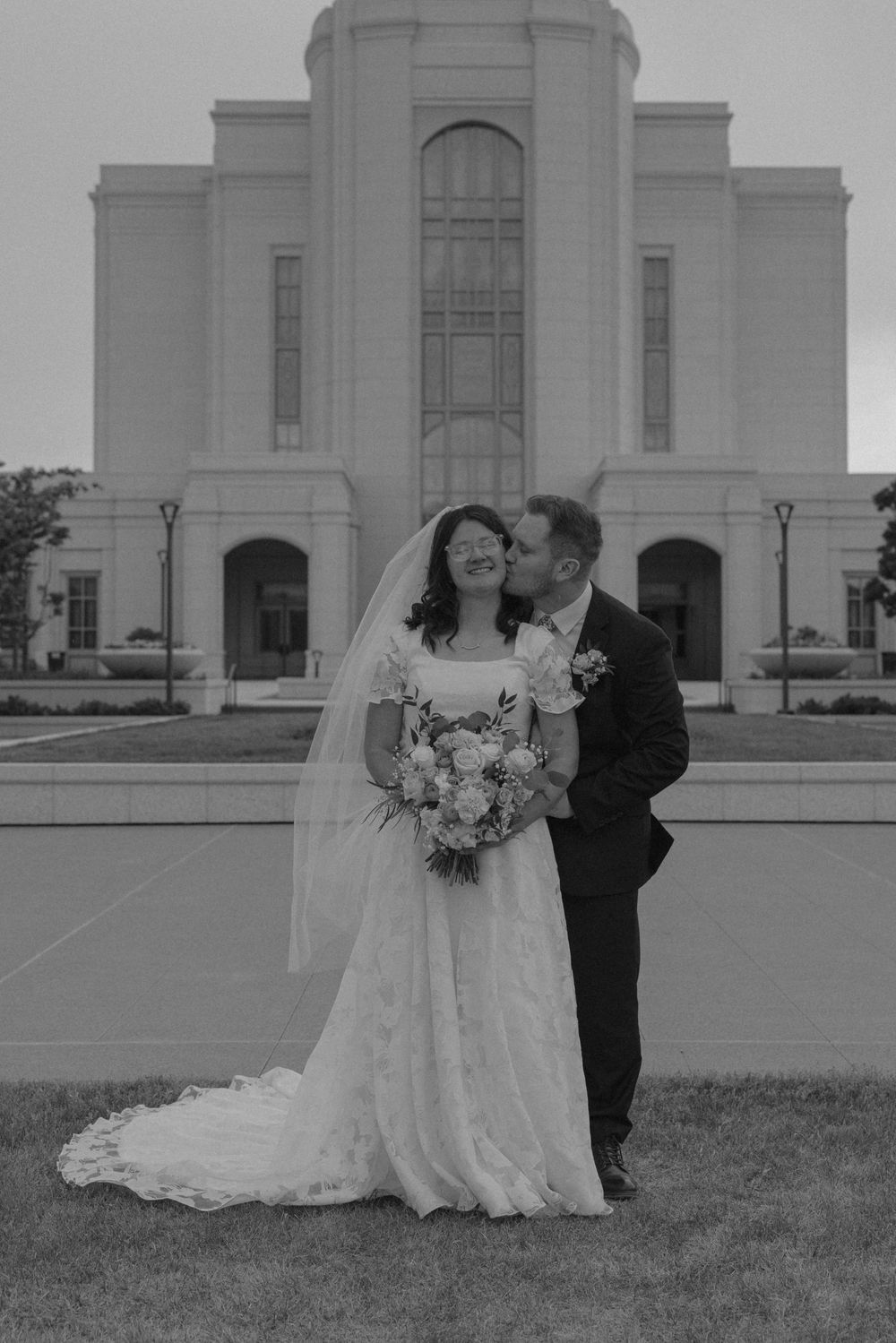 Groom kisses bride on cheek as she smiles holding bouquet in black and white temple wedding portrait.