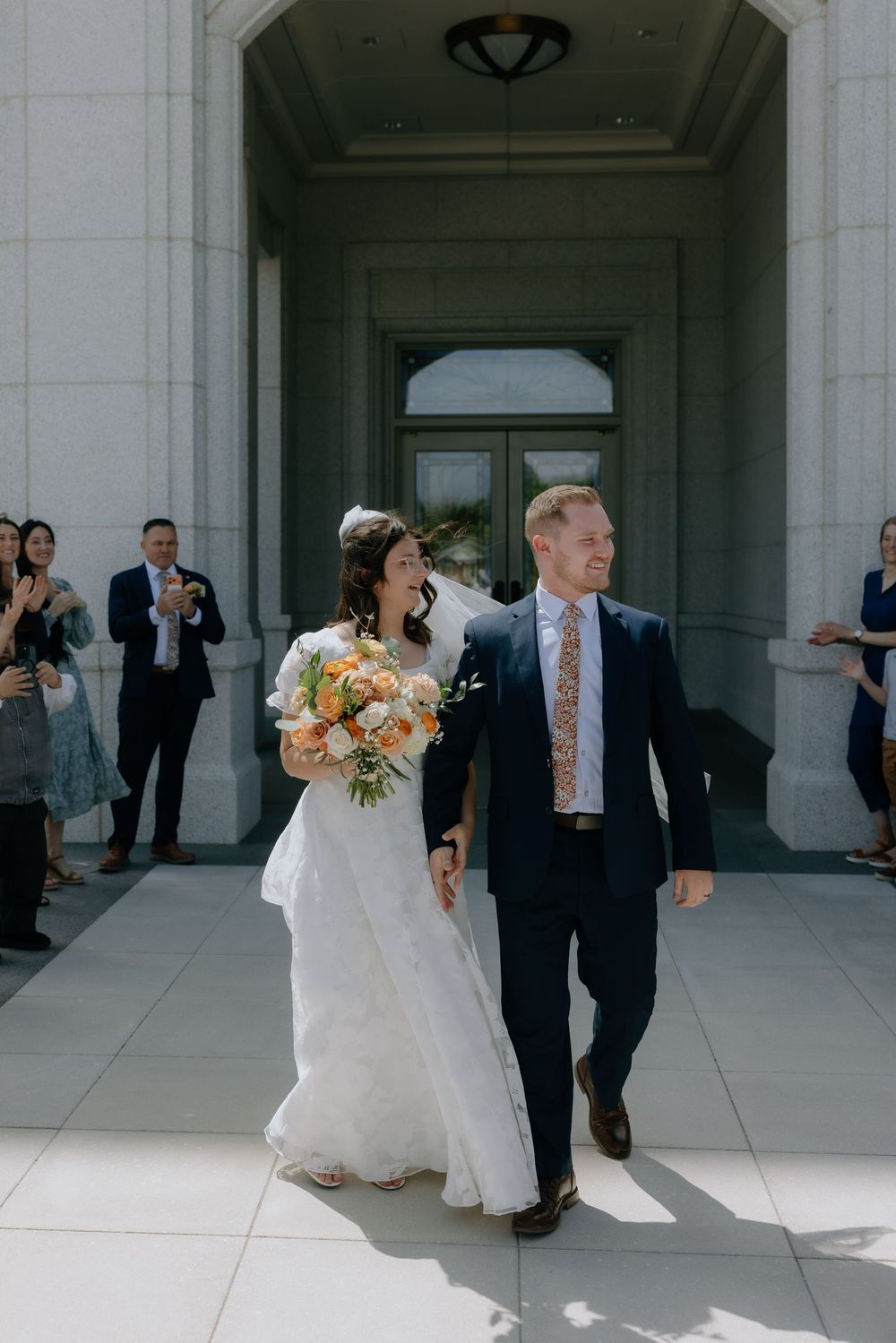 Bride and groom exit wedding venue smiling, bride holds peach bouquet, guests applaud in background.