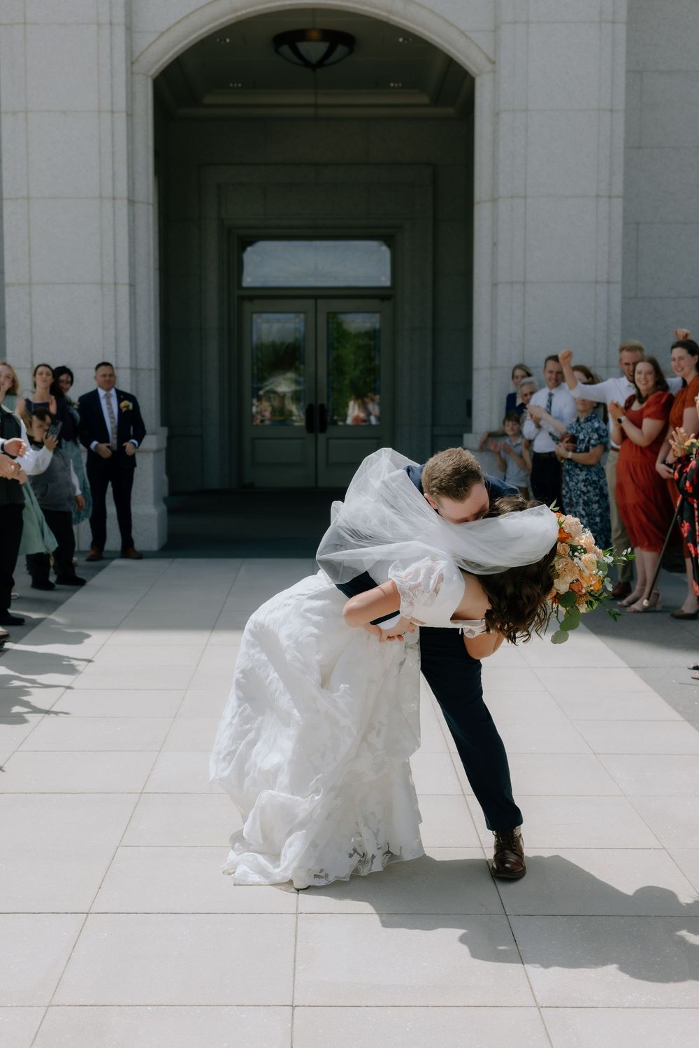 Groom dips bride in romantic kiss outside temple as wedding guests look on in sunny outdoor ceremony.