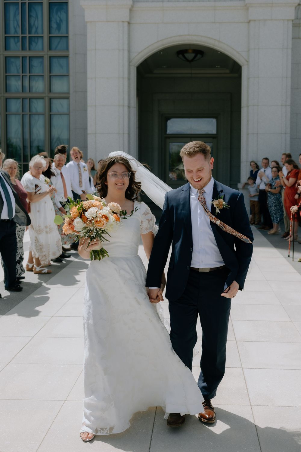 Smiling bride and groom walk hand in hand from temple doors as guests celebrate their wedding day.