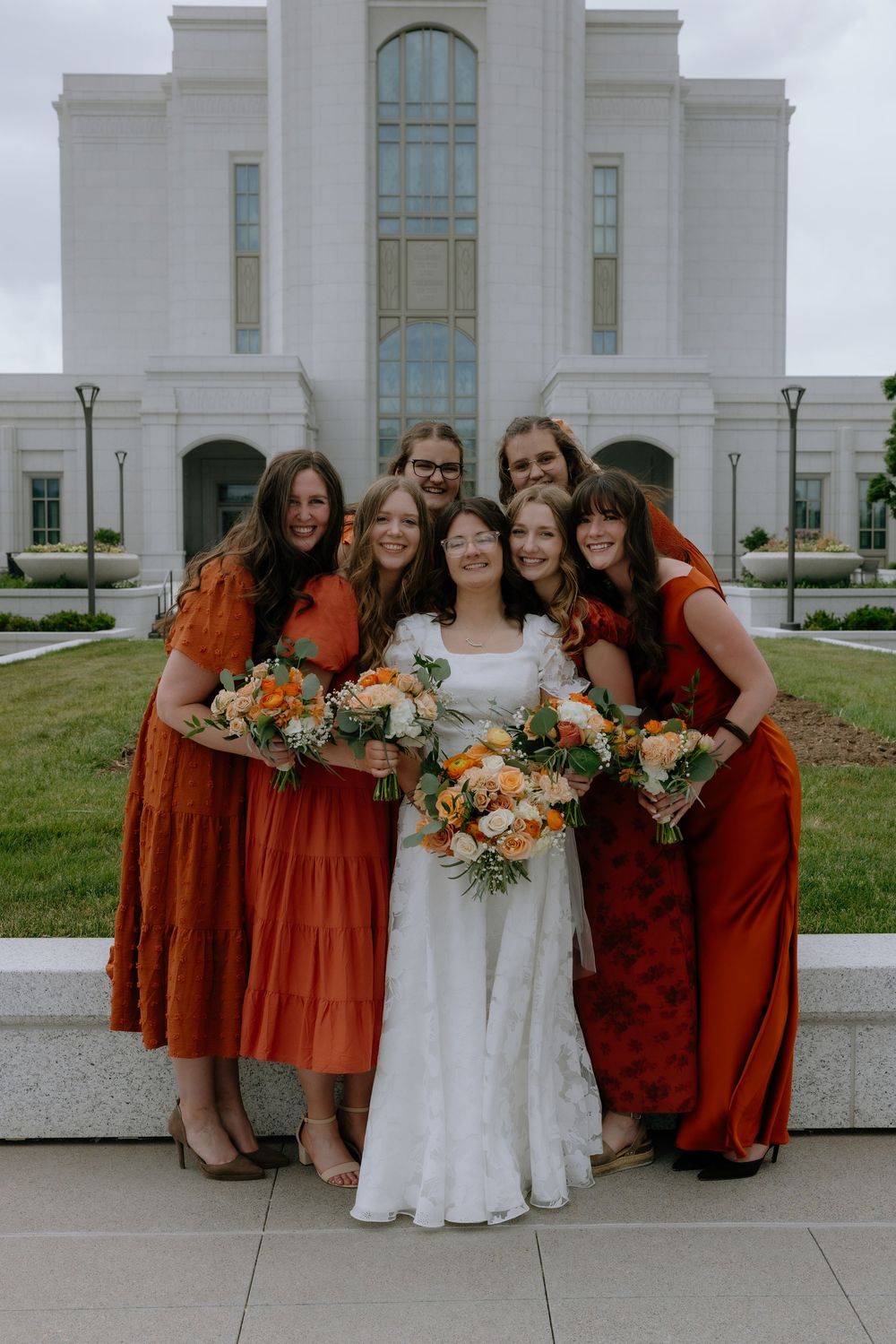 Bride in white lace gown poses with six bridesmaids in rust orange dresses holding bouquets outside a white temple.