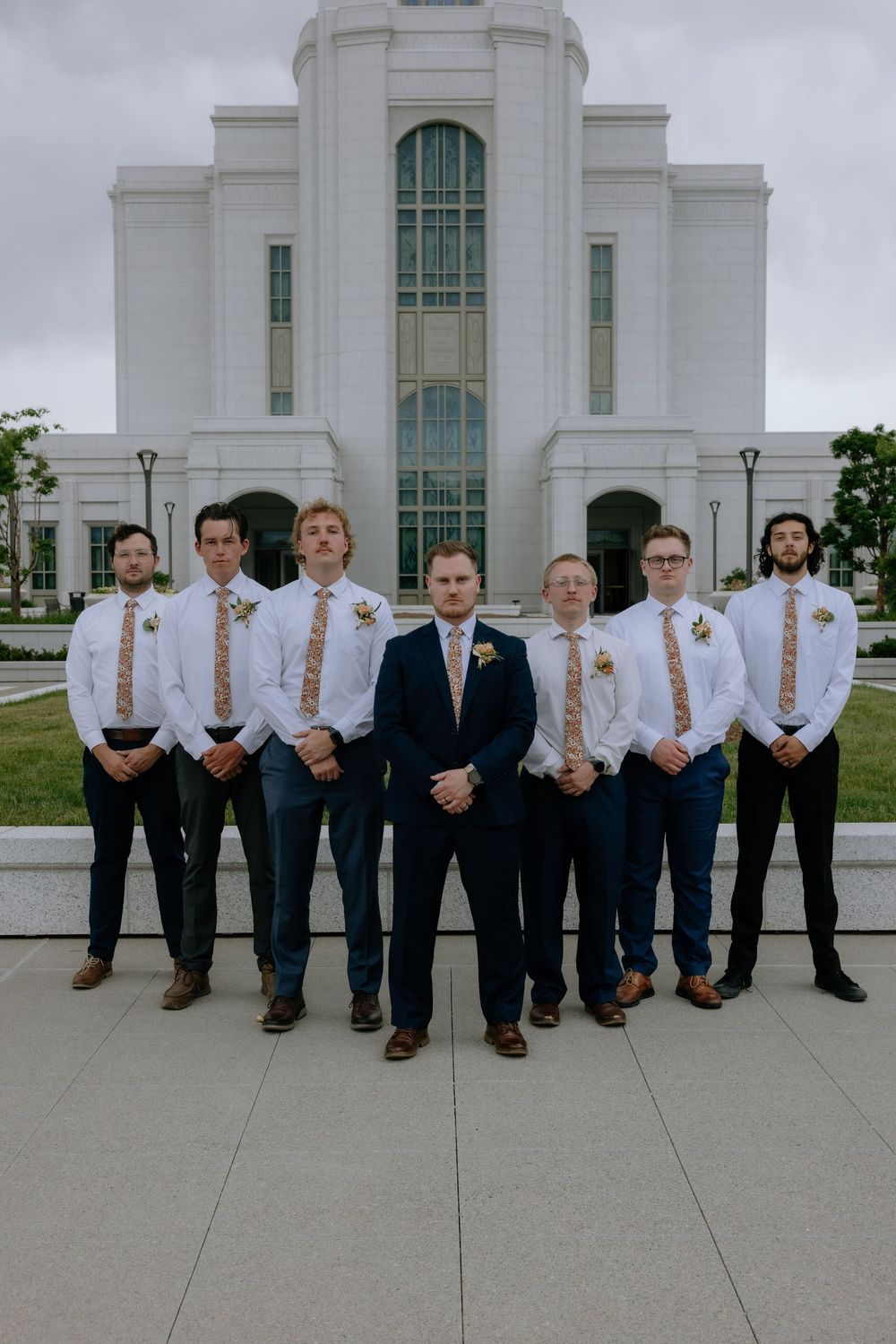 Groom and groomsmen in white shirts and floral ties pose outside a white LDS temple on a wedding day.