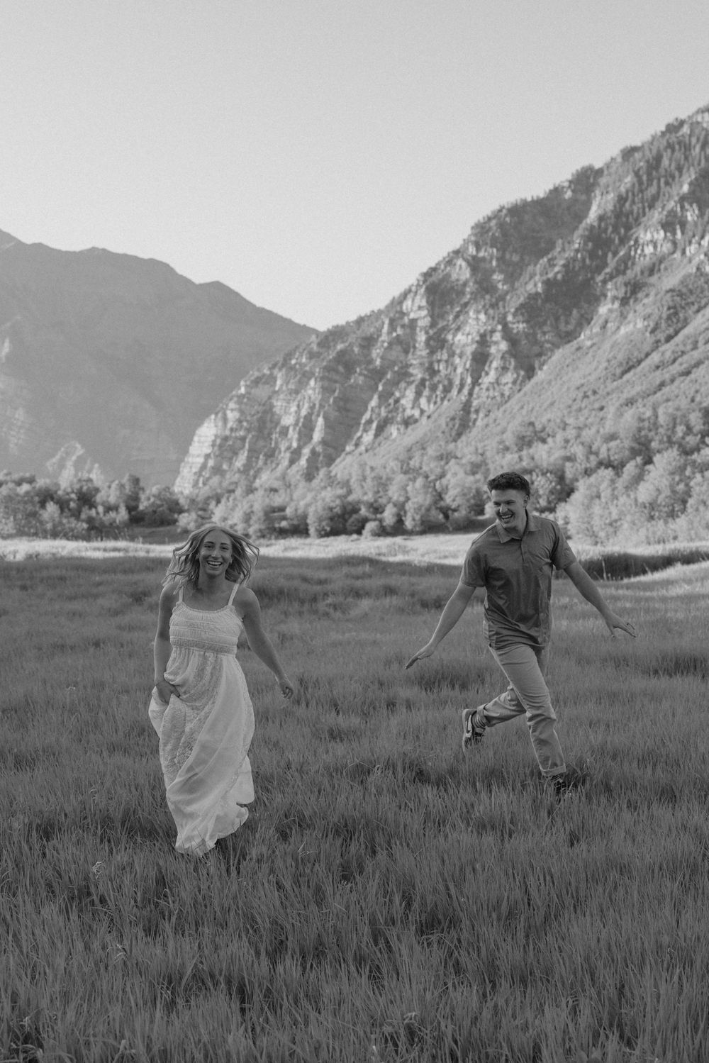 Black and white photo of a joyful couple running through a mountain meadow, woman in white dress, man in casual attire.
