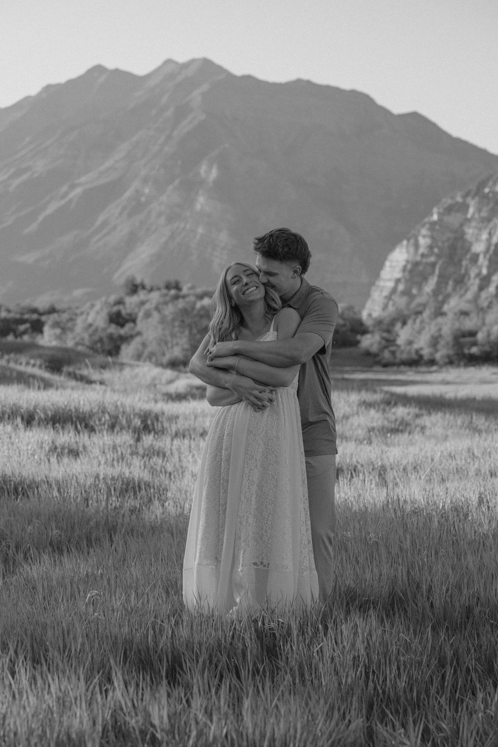 Black and white photo of a couple embracing in a field with mountains in the background.