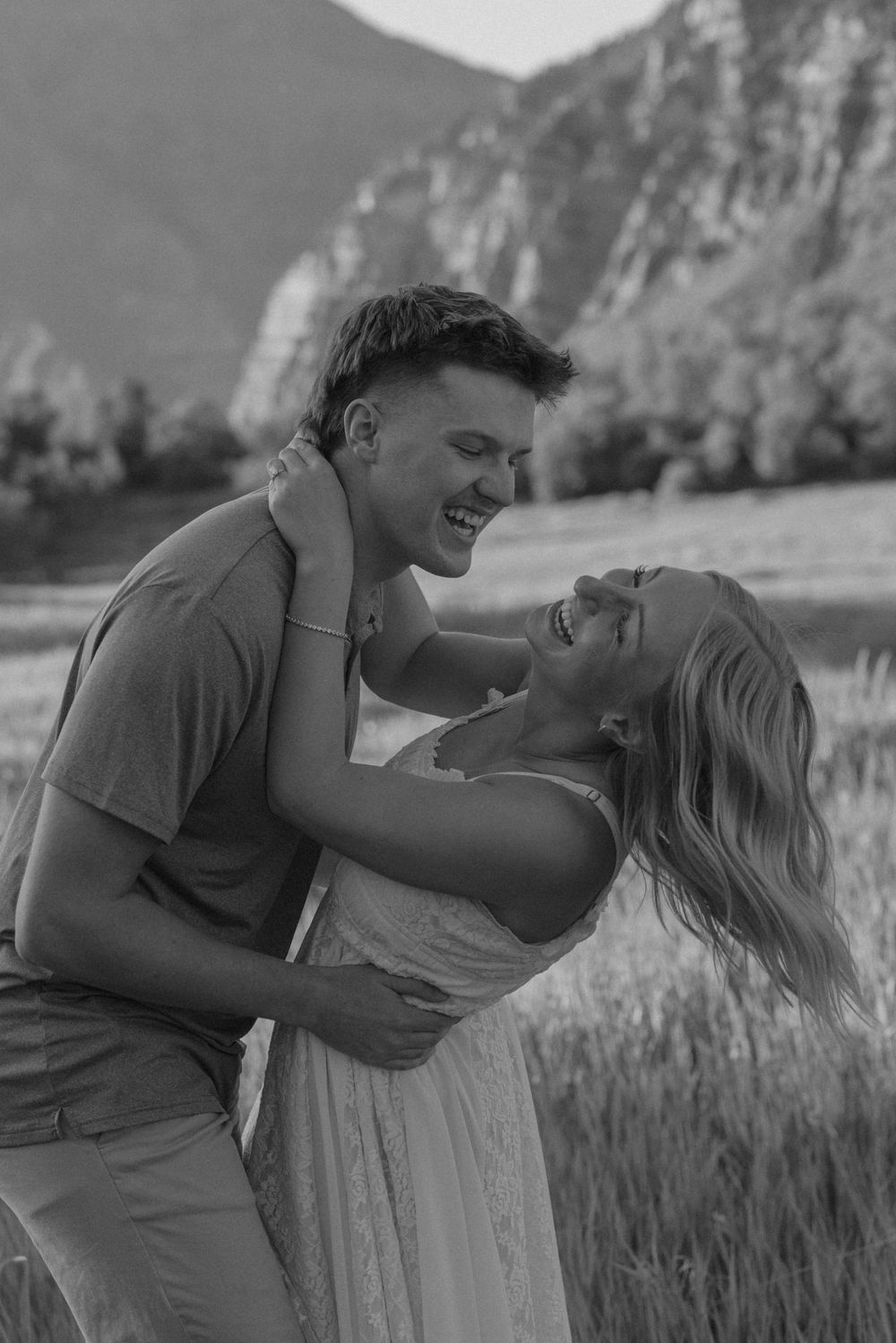 Black and white photo of a laughing couple embracing in a field with mountains in the background.