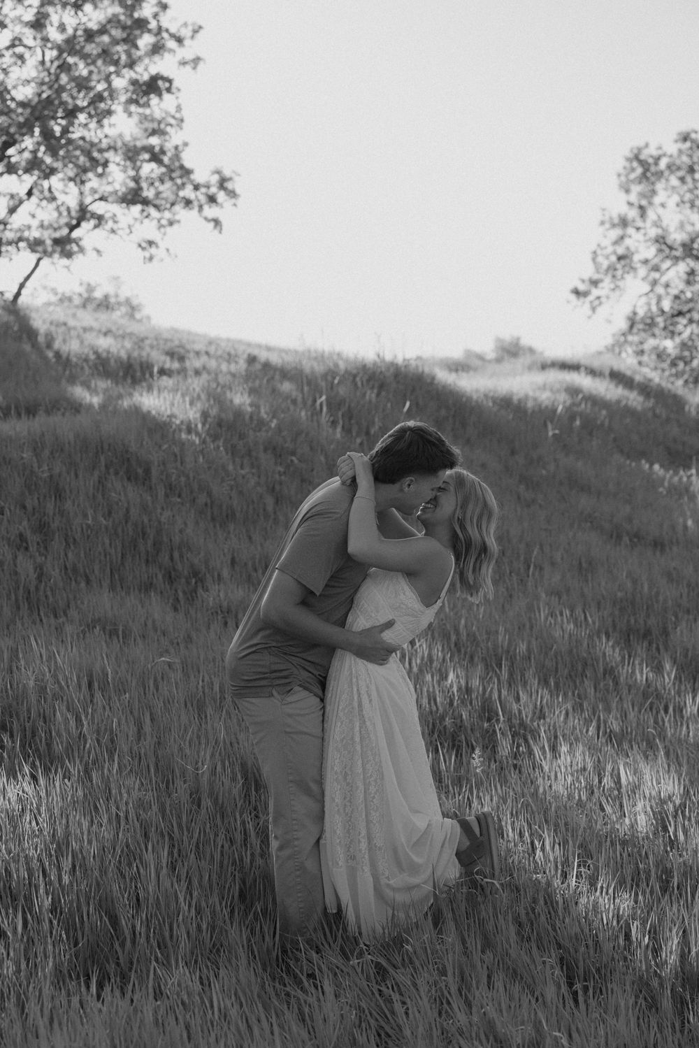 Black and white romantic photo of couple embracing on a grassy hillside with tall grass surrounding them.