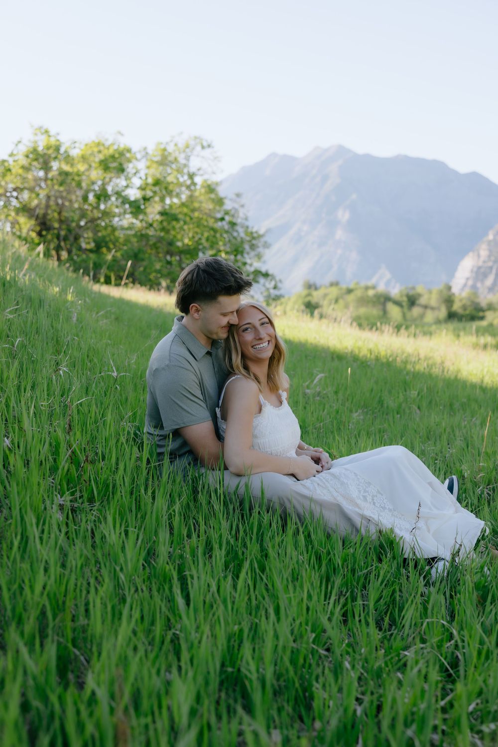 A couple sits together in lush green grass with a mountain backdrop during golden hour.