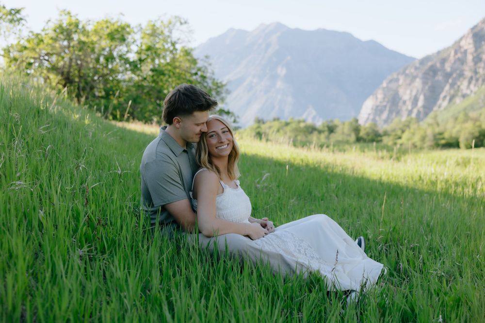 Couple sitting together in lush green meadow with mountain backdrop, sharing a romantic moment outdoors.