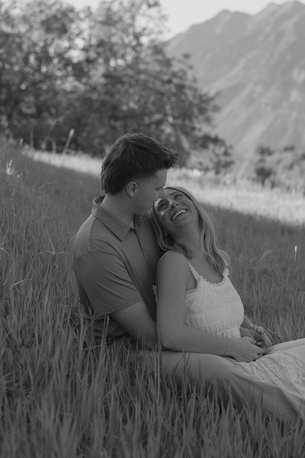 Couple sitting in tall grass laughing together in a black and white photo.