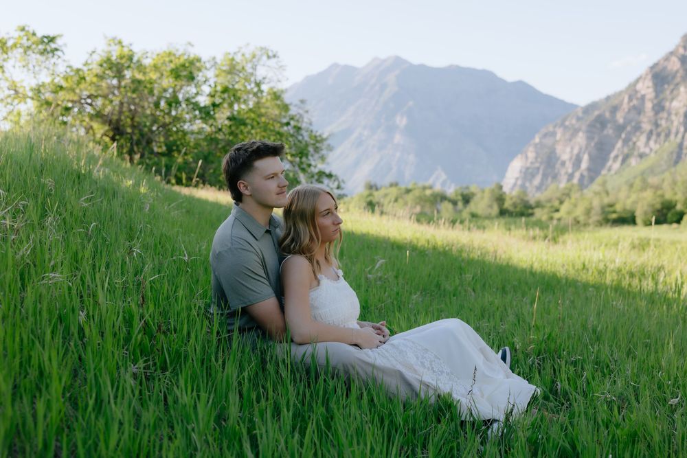 Couple sitting together in lush green meadow with mountain backdrop during golden hour engagement photo session.