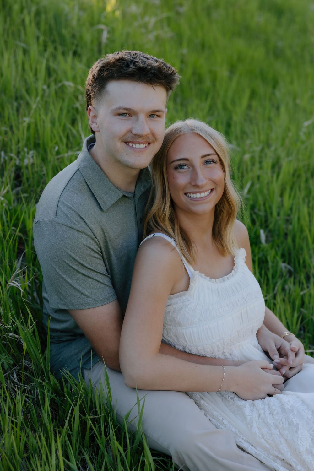 Smiling couple sitting together in lush green grass during golden hour engagement photos.