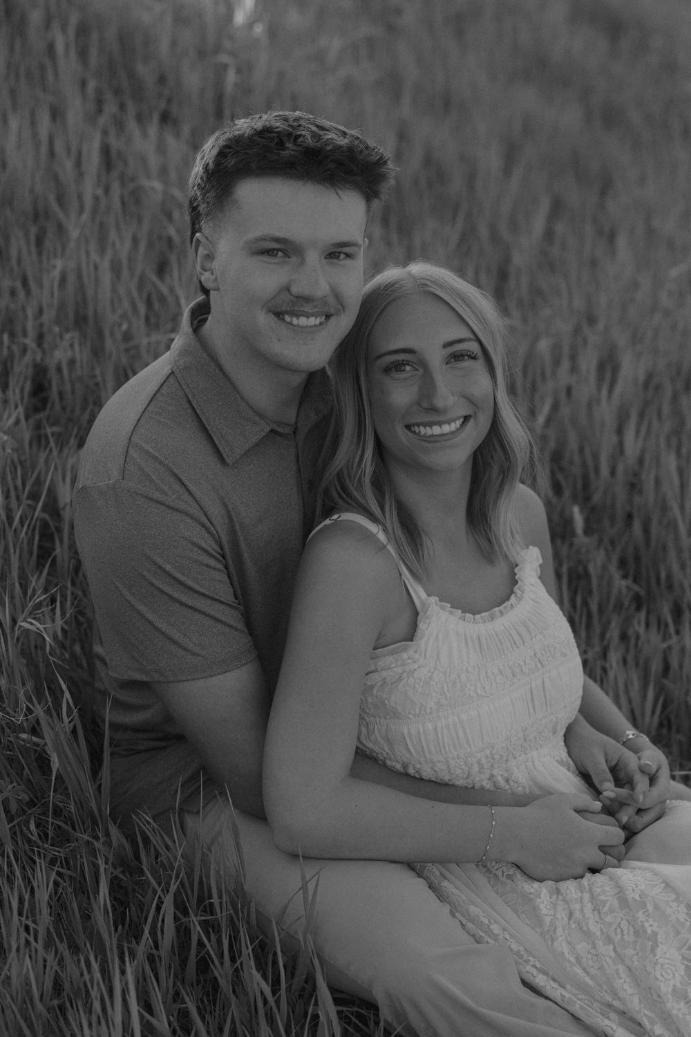 Black and white portrait of smiling couple sitting together in a tall grass field, looking at camera.