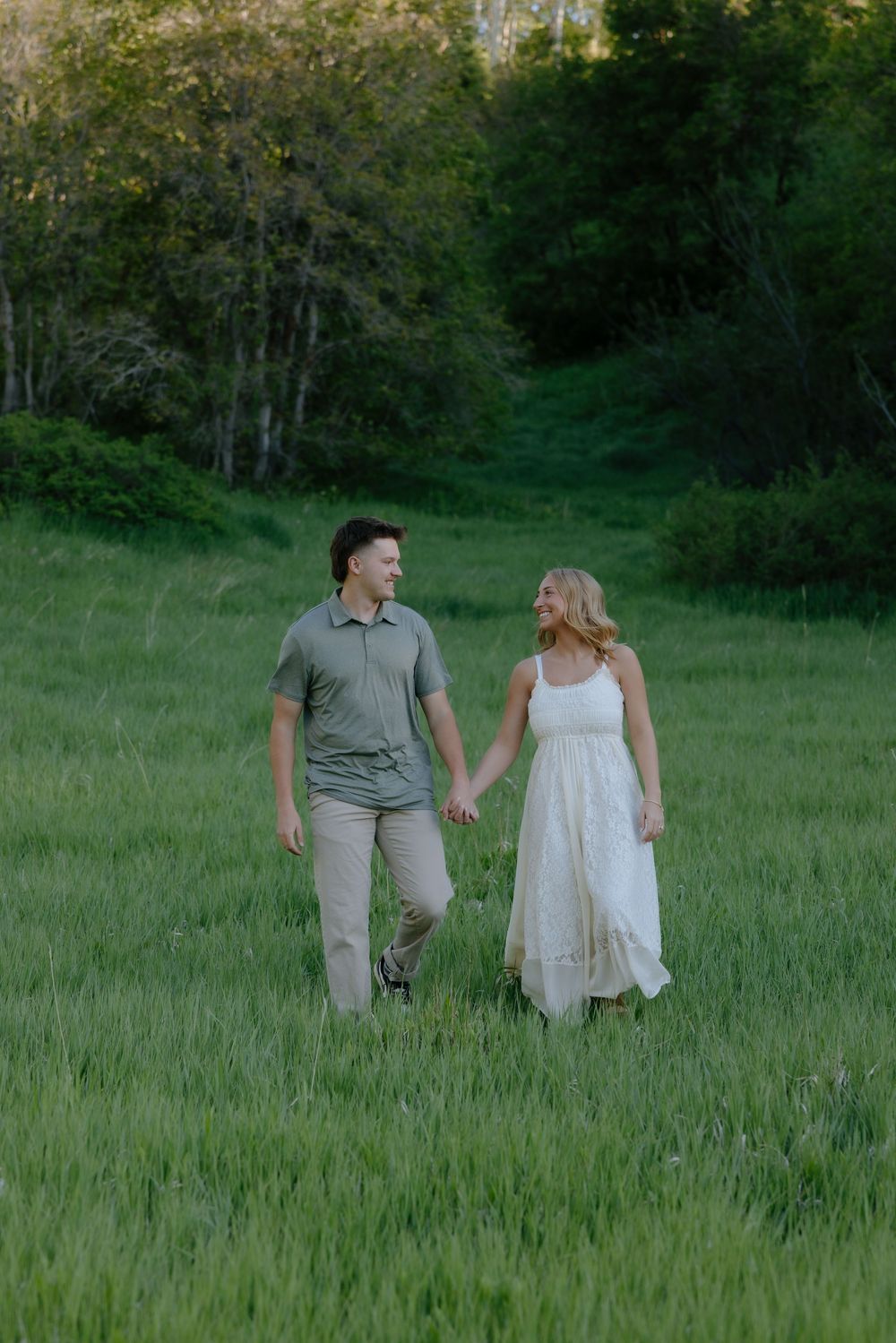 Couple holding hands and smiling while walking through a lush green meadow at dusk.