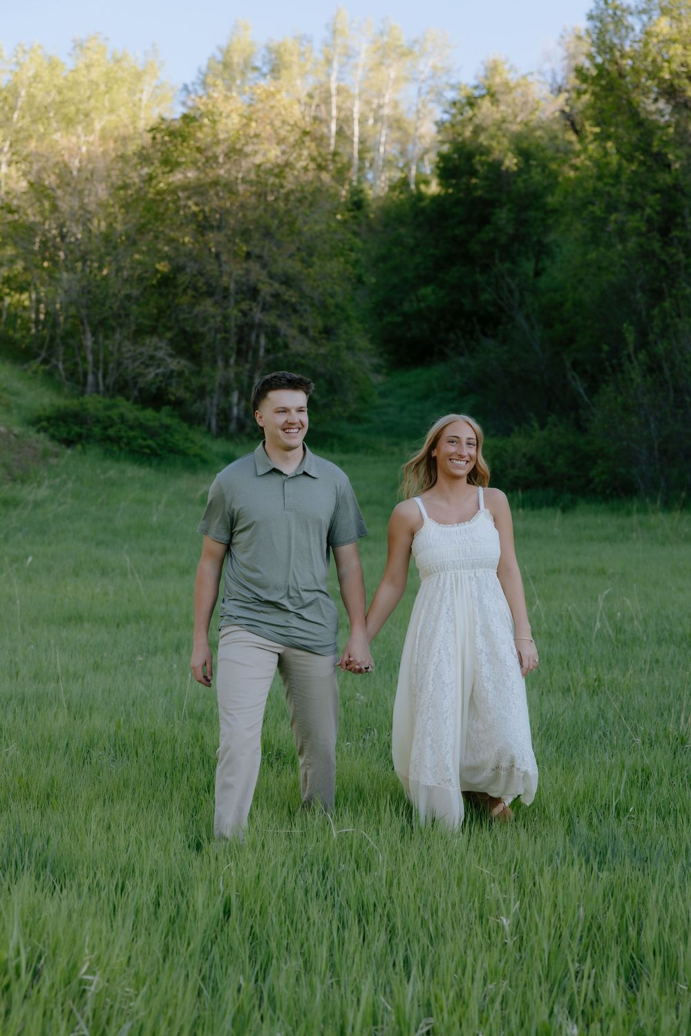 A smiling couple holds hands while walking through a green meadow with trees behind them.