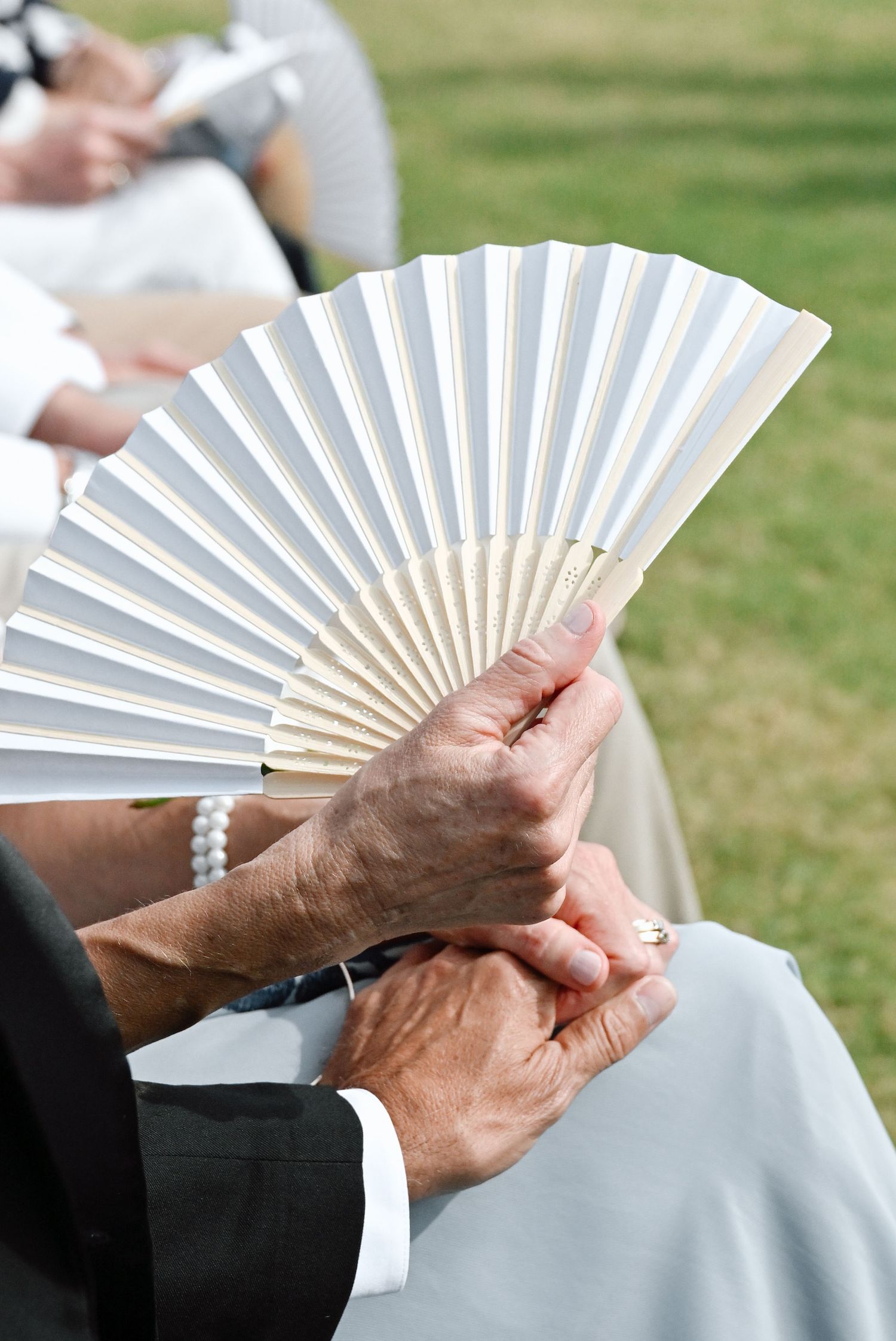 Elderly hands holding a white paper fan at an outdoor wedding ceremony on a sunny day.