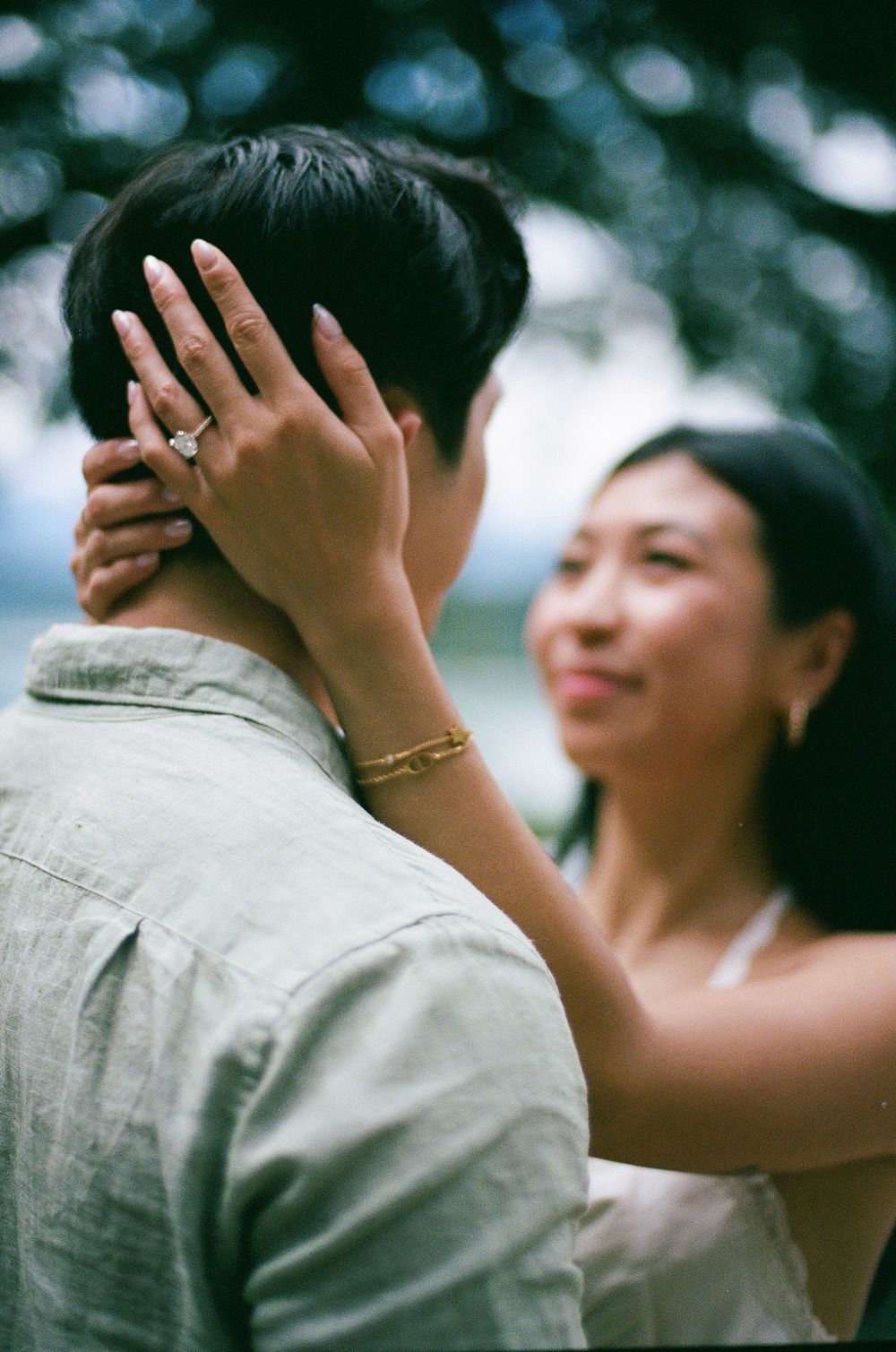 Couple sharing a tender moment, woman with diamond ring and gold bracelet gently holding mans face outdoors.