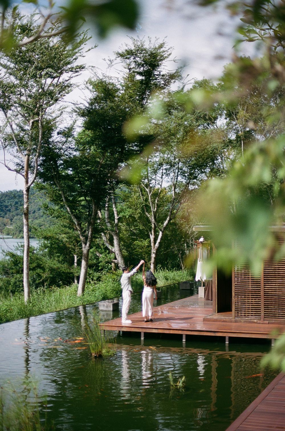 Two people stand on a wooden deck beside a serene natural pond surrounded by lush green trees and tropical foliage.