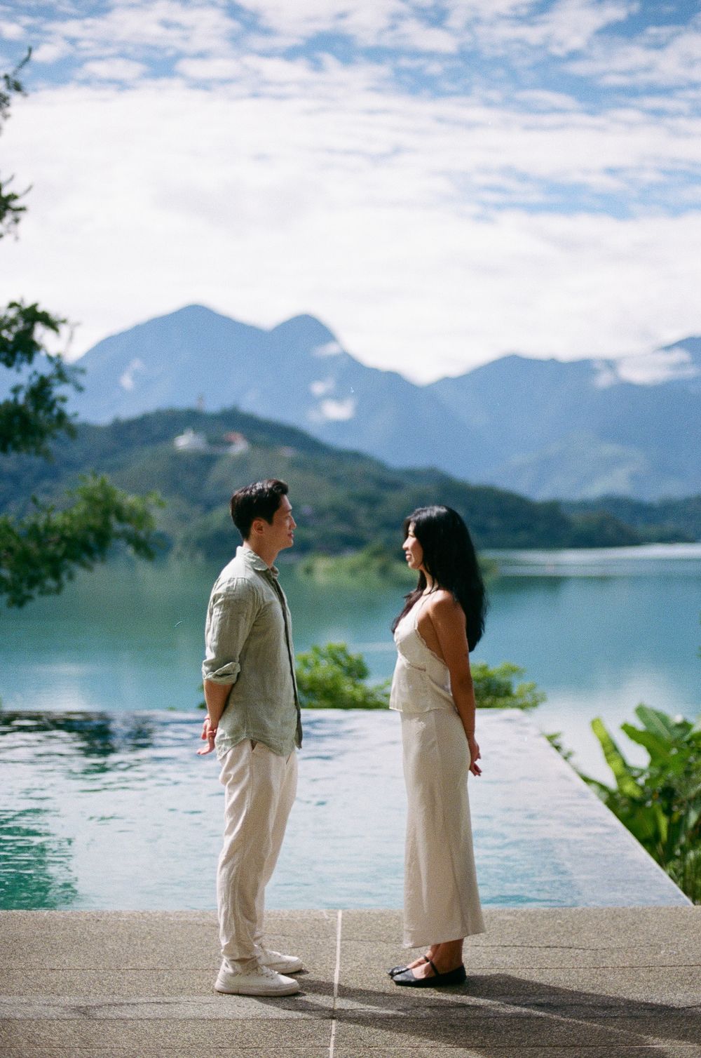 Couple facing each other by an infinity pool with turquoise lake and misty mountains in the background.