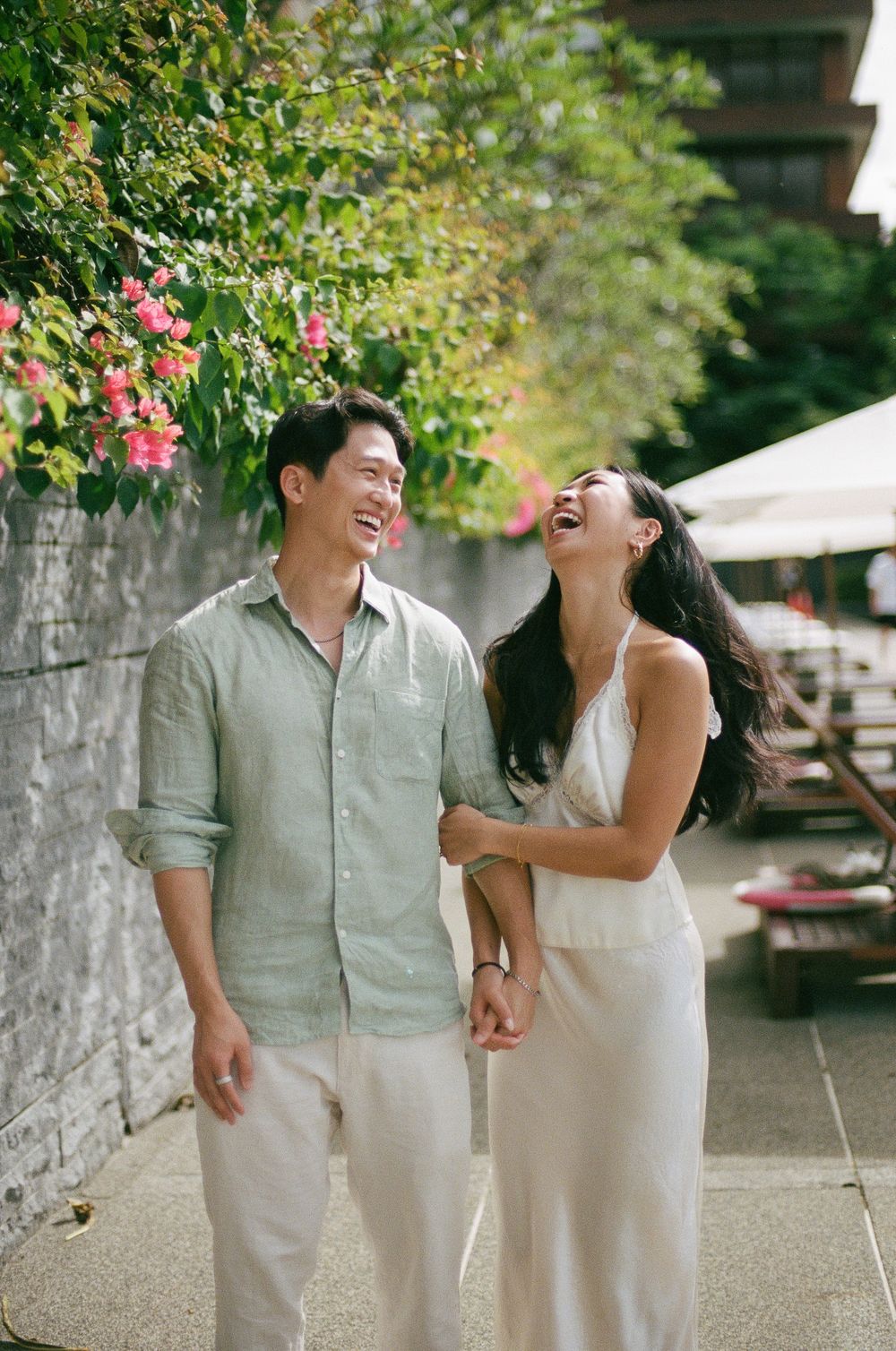 A joyful Asian couple laughs together while walking along a garden path lined with pink flowers.