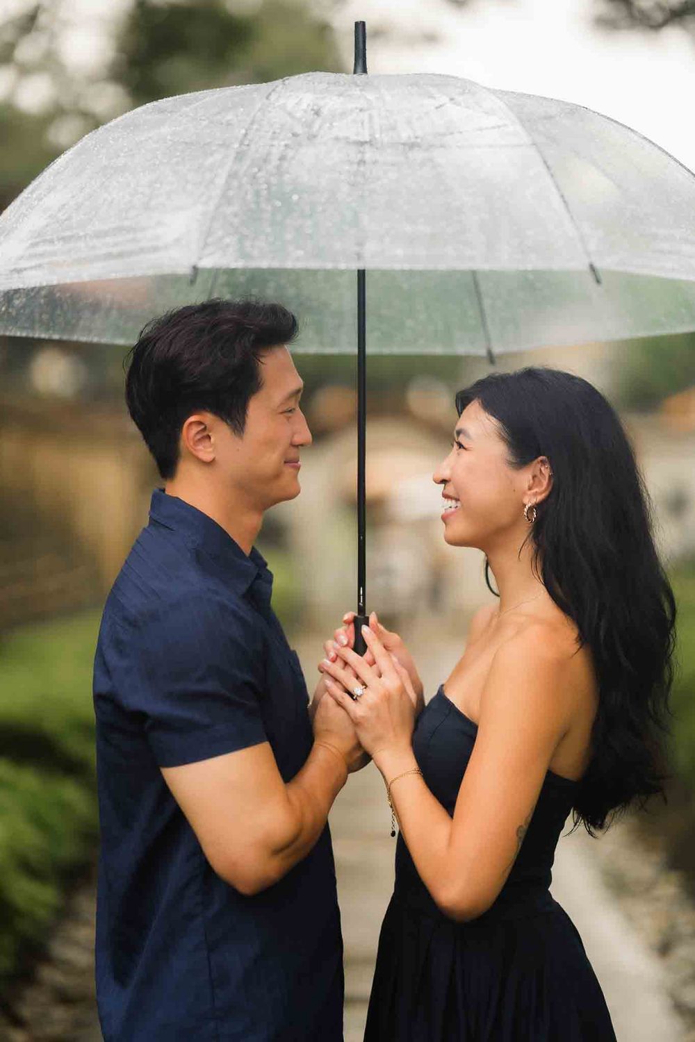 Couple sharing a clear umbrella in the rain, smiling at each other in a romantic outdoor setting.