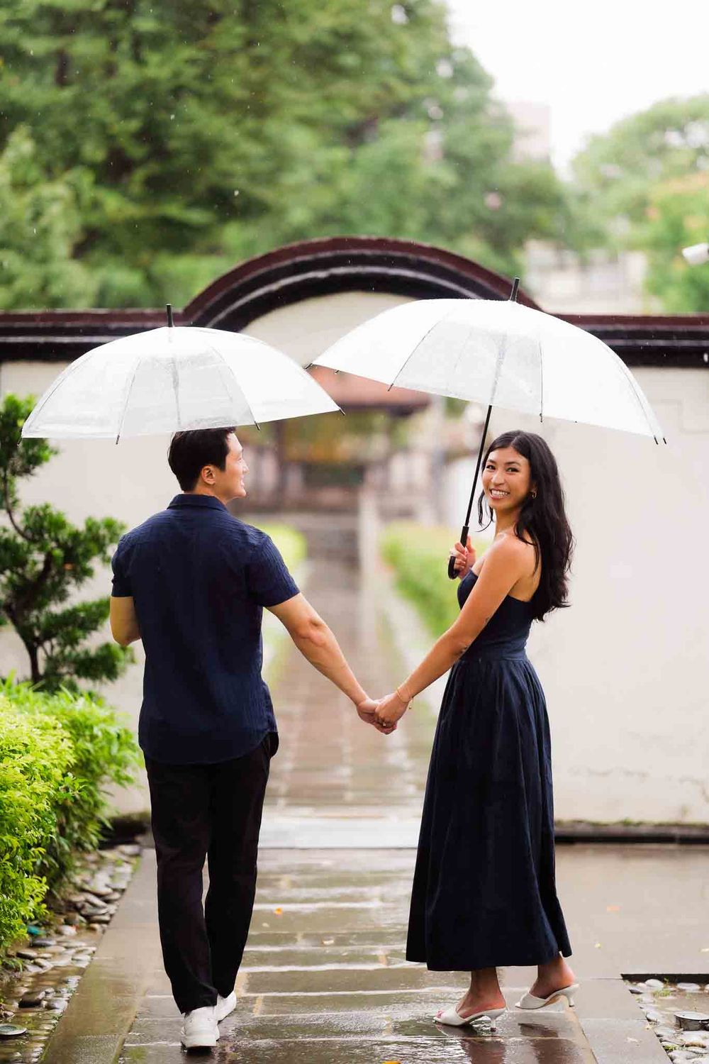 Couple holding hands and clear umbrellas in the rain at a scenic Asian garden pathway.