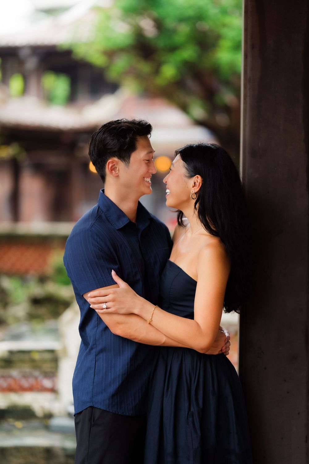 A smiling Asian couple in matching navy outfits share a romantic embrace outdoors near a wooden pillar.