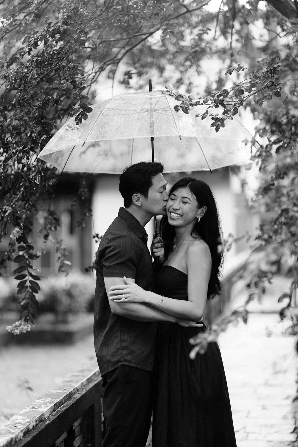 Black and white photo of a couple sharing a romantic moment under a clear umbrella in the rain.