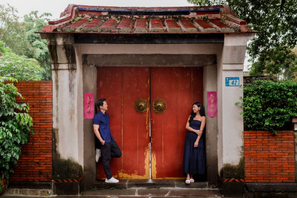 Couple posing on either side of a red traditional Chinese gate with brick walls and lush greenery.