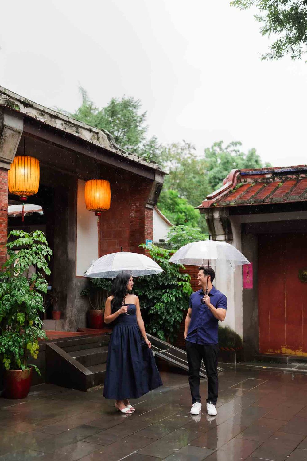 Couple holding clear umbrellas in the rain at a traditional Asian courtyard with red lanterns and tiled roofs.